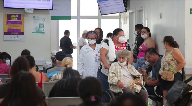 A foto mostra Mônica de pé em meio a uma área de espera da unidade de saída, cercada de dezenas de pacientes 